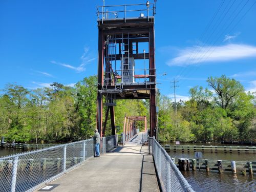 A tall bridge structure over a calm river, surrounded by greenery and blue skies, with a person standing nearby.