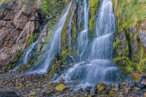 A serene waterfall cascading over moss-covered rocks, surrounded by lush greenery and smooth pebbles below.