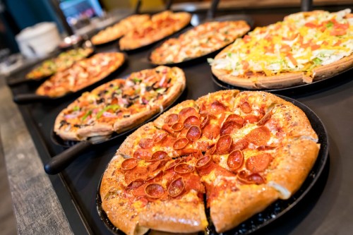 A variety of pizzas on display, including pepperoni, veggie, and taco-style, on black serving trays.