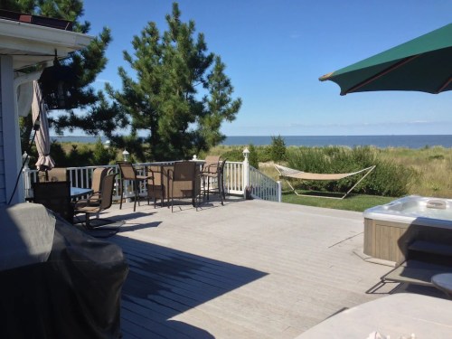 A sunny outdoor deck with seating, a hammock, and a hot tub, overlooking a calm body of water and green landscape.