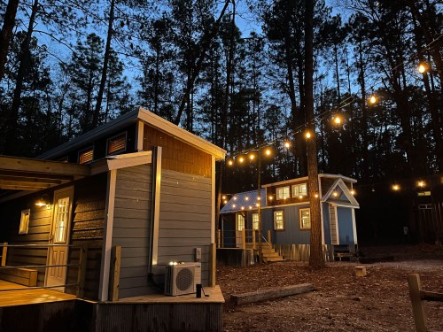 Two cozy cabins illuminated by string lights, surrounded by tall trees at dusk.