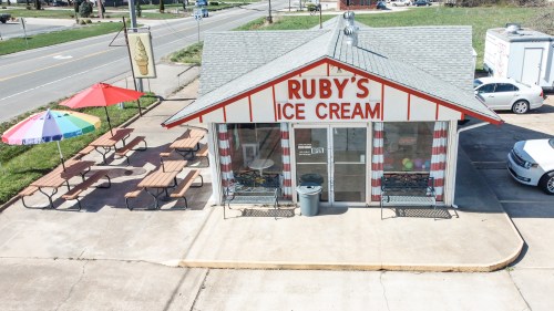 A colorful ice cream shop with a sign reading "Ruby's Ice Cream," featuring outdoor seating and umbrellas.