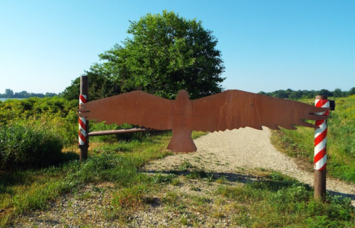 A metal sculpture of an eagle marks the entrance to a gravel path surrounded by greenery and blue sky.