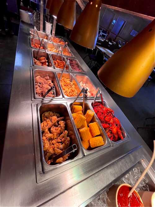 A buffet table with various dishes, including fried chicken, corn, seafood, and sauces in metal trays.