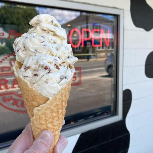A hand holding a large ice cream cone in front of a shop window with a neon "OPEN" sign.