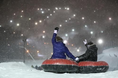 Two people sitting on snow tubes, waving in a snowy landscape with lights twinkling in the background.