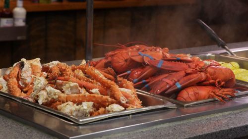 A seafood display featuring cooked lobster and crab legs in metal trays, with steam rising in the background.