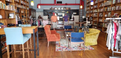 A cozy bookstore with wooden shelves filled with books, colorful seating, and clothing displays.