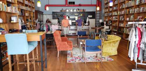 A cozy bookstore with wooden shelves filled with books, colorful seating, and clothing displays.