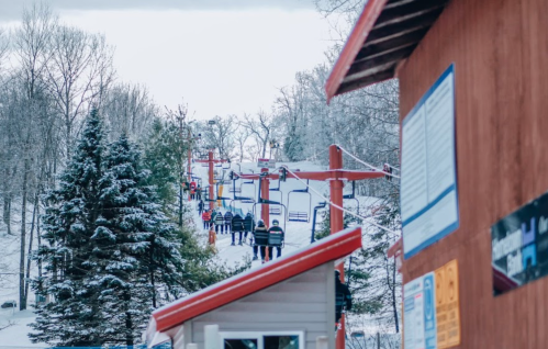 Ski lift with people ascending a snowy hill, surrounded by trees and a wooden structure in the foreground.