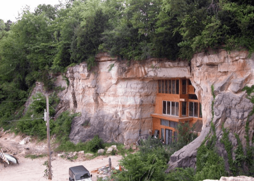 A modern house built into a rocky cliff, surrounded by greenery and a gravel area with a parked vehicle.