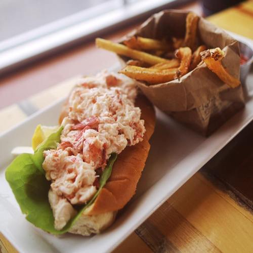 A lobster roll with lettuce on a bun, served with a side of fries in a paper cup.