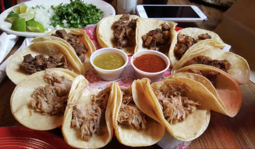 A colorful platter of tacos with various meats, accompanied by sauces, lime, and fresh cilantro on the side.