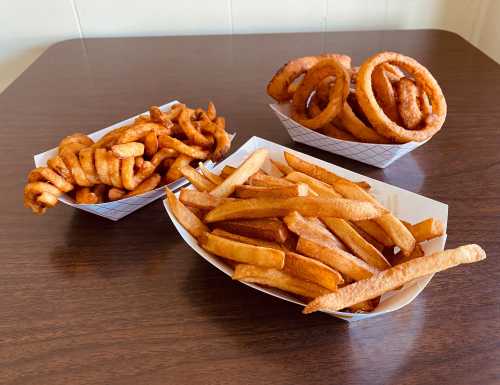 Three types of fried snacks on a wooden table: curly fries, onion rings, and straight-cut fries in paper trays.