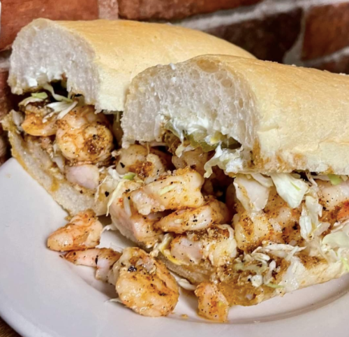 A close-up of a shrimp sandwich with lettuce on a plate, served on a soft white roll against a brick background.