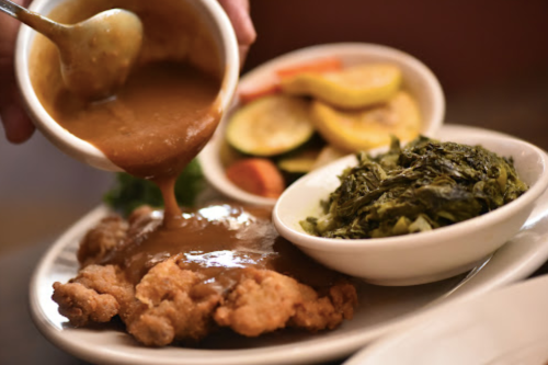 A plate of fried chicken topped with gravy, served with greens and a side of mixed vegetables.