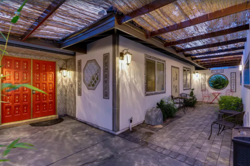 A cozy outdoor corridor with a red door, stone accents, and seating under a thatched roof, illuminated at dusk.