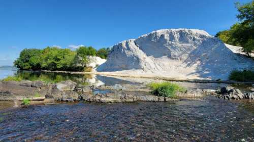 A sandy hill beside a calm river, surrounded by greenery and a clear blue sky. Reflections shimmer on the water's surface.