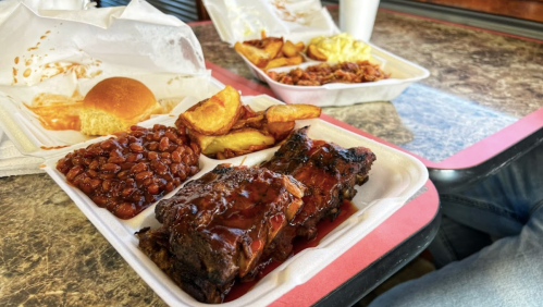 A takeout meal featuring ribs with barbecue sauce, baked beans, potato wedges, a roll, and a side of macaroni and cheese.