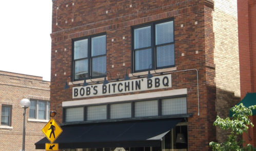 Sign for Bob's Bitchin' BBQ on a brick building, featuring large black letters and a pedestrian crossing sign nearby.