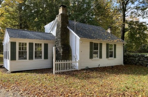 A charming white cottage with green shutters, a stone chimney, and a picket fence, surrounded by autumn foliage.
