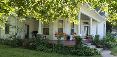 A charming white house with a porch, surrounded by greenery and trees, featuring a swing and a grill.