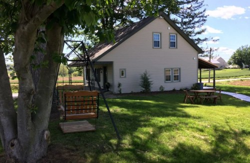 A cozy two-story house with a porch, swing, and green lawn, surrounded by trees and open fields under a blue sky.