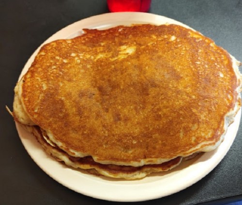 A stack of golden-brown pancakes on a white plate, resting on a dark surface.