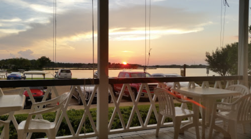 Sunset view over a calm lake from a porch with white chairs and parked cars in the foreground.