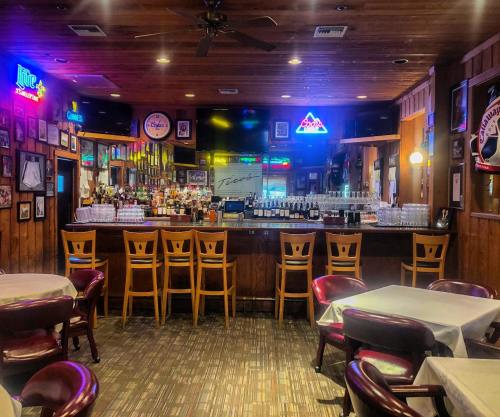 A cozy bar area with wooden decor, empty stools, and colorful neon signs, set in a casual dining space.