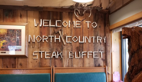 Wooden sign reading "Welcome to North Country Steak Buffet" on a rustic wall with a bear sculpture nearby.