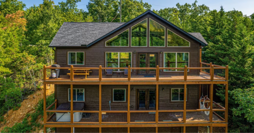 A modern two-story cabin surrounded by trees, featuring large windows and a spacious wooden deck.