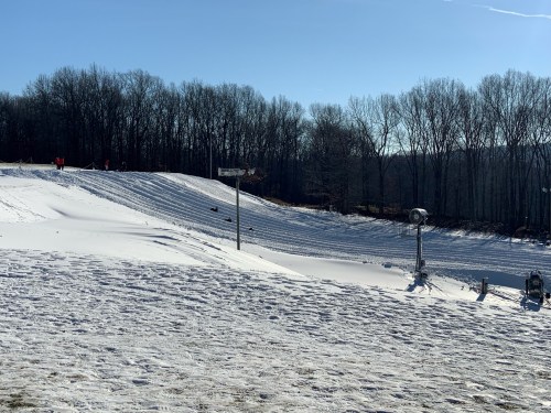 A snowy landscape with a hill, trees in the background, and a clear blue sky. People are visible on the slope.