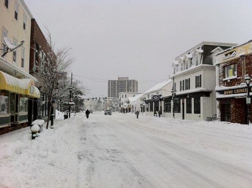Snow-covered street with buildings on either side, people walking, and a tall building in the background.