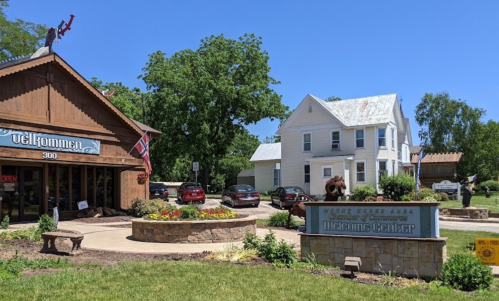 A charming town scene featuring a wooden building, a welcome sign, and a white house surrounded by greenery and parked cars.