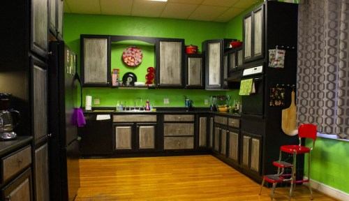 A modern kitchen with green walls, black cabinets, and wooden floors, featuring a red chair and various kitchen appliances.