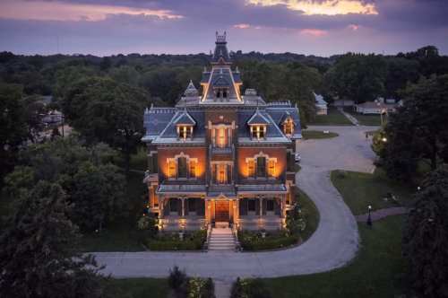 Aerial view of a grand, illuminated Victorian mansion surrounded by trees at dusk.