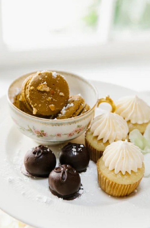 A plate of assorted desserts: macarons, chocolate truffles, and frosted cupcakes, with a floral bowl in the background.