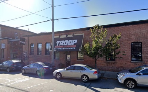 Exterior of a brick building with a large "TROOP" sign, surrounded by parked cars on a sunny day.