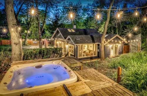 A cozy cabin with a hot tub, surrounded by trees and string lights, set in a serene outdoor environment at dusk.