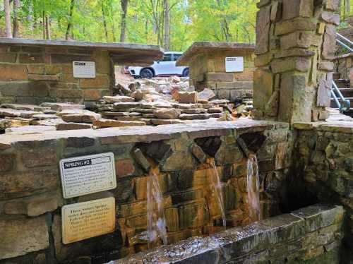 A stone structure with flowing water, surrounded by trees, with a vehicle visible in the background.