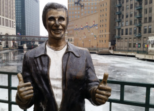 A bronze statue of a smiling man giving a thumbs-up, with a frozen river and city buildings in the background.
