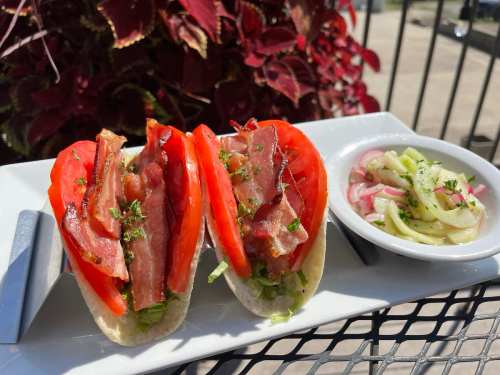 Two tacos filled with lettuce, tomato, and bacon, served with a side of cucumber and onion salad.