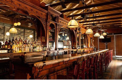 A vintage-style bar with wooden decor, illuminated by pendant lights, showcasing a variety of bottles on shelves.