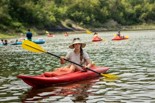 A person in a red kayak paddles on a calm river, surrounded by others enjoying kayaking on a sunny day.