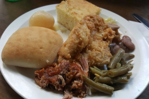 A plate of food featuring fried chicken, cornbread, green beans, beans, a roll, and a fruit cup.