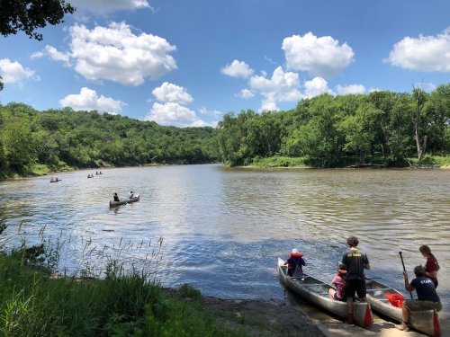 A serene river scene with people canoeing and enjoying a sunny day surrounded by lush greenery and blue skies.