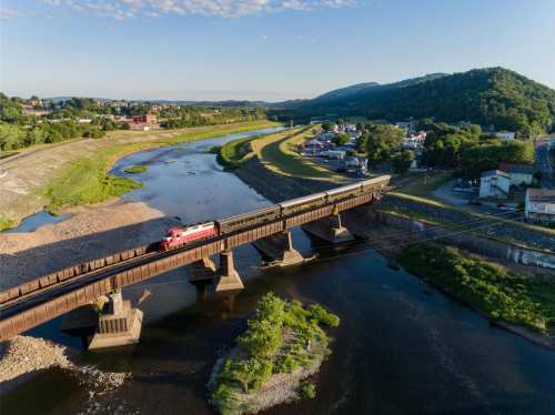 A train crosses a bridge over a river, surrounded by green hills and a small town in the background.