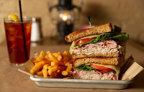 A stacked sandwich with turkey, lettuce, and tomato, served with curly fries and a drink in the background.