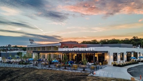 A brewery building with outdoor seating, surrounded by greenery, under a colorful sunset sky.